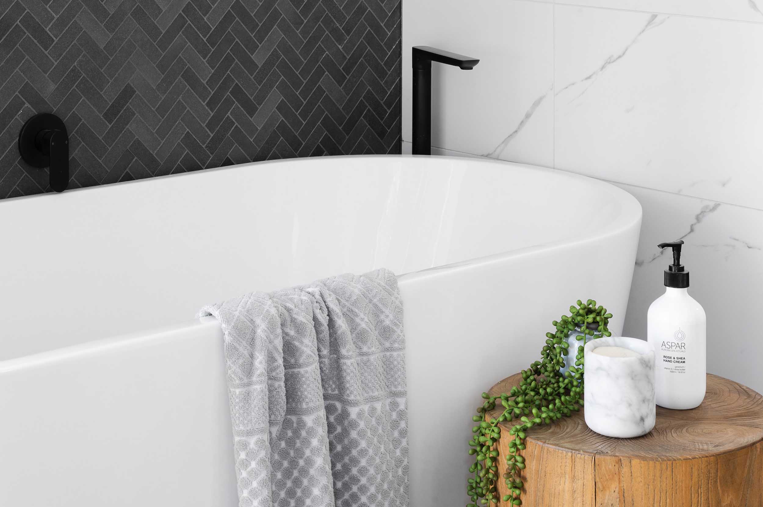Nyrenoverat badrum med badkar, svart & marmor kakel och en trästubbe med en växt och doftljus. Tony Söderström Bygg & Kakel